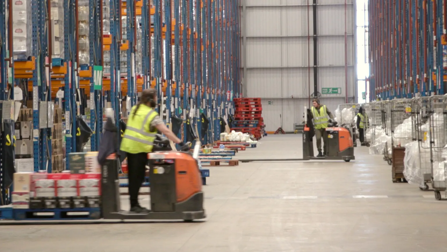 Wide-angle view of a high-bay warehouse with workers operating electric pallet trucks along a central aisle, flanked by tall racking and staged pallets. Ideal for illustrating independent supply chain integrity audits, goods receiving verification, pick accuracy checks, and logistics performance assurance in regulated retail environments. Wide-angle view of a high-bay warehouse with workers operating electric pallet trucks along a central aisle, flanked by tall racking and staged pallets. Ideal for illustrating independent supply chain integrity audits, goods receiving verification, pick accuracy checks, and logistics performance assurance in regulated retail environments.